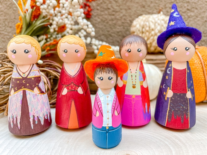 Op de afbeelding: Vijf houten pegdolle met beschilderde gezichten en kleding. De poppen zijn gekleed in verschillende outfits, waaronder een rode jurk, een paarse robe, een roze jurk, een blauw shirt en jeans, en een bruine jurk met een gouden ketting. De poppen staan op een houten oppervlak.