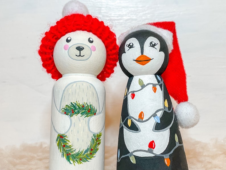 May include: Two hand-painted wooden peg dolls dressed in festive attire. The first doll is a polar bear wearing a red knit hat and holding a green wreath. The second doll is a penguin wearing a red Santa hat and decorated with colorful lights.