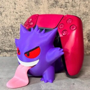 May include: A purple Gengar-shaped controller holder with a pink tongue, red eyes, and a mischievous grin. A red and black game controller is behind the Gengar figure. The holder is designed to hold a game controller.