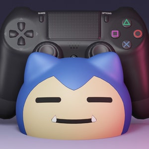 Op de afbeelding: Een zwarte PlayStation 4-controller staat rechtop met een blauw en geel cartoonfiguur ervoor. Het figuur heeft een brede glimlach en twee grote hoektanden.