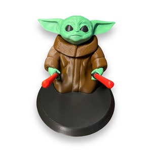 May include: A green and brown figurine of a character from the Star Wars franchise, holding two red lightsabers. The figurine is standing on a black circular base.