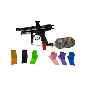 Puede incluir: Una pistola de paintball negra con un tanque de camuflaje se muestra en un soporte rojo. La pistola está apoyada en un soporte de varios colores: amarillo, negro, verde, rosa, morado y azul.