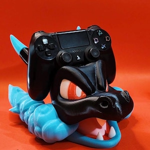 Peut inclure: Un contrôleur de jeu noir est posé sur un support imprimé en 3D sur le thème de Lucario. Le support est noir avec des yeux rouges et une queue et une base bleues et pointues. Le fond est orange uni.
