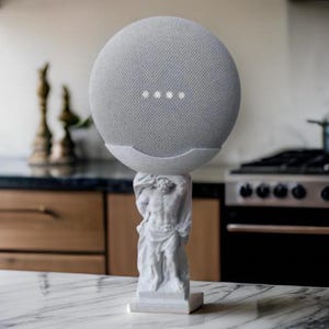 May include: A grey smart speaker is displayed on a white statue of a classical male figure. The speaker has four white indicator lights. The statue is on a white marble countertop in a kitchen.