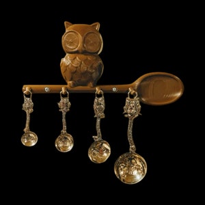 Puede incluir: Una figurita de búho marrón posada sobre una cuchara de madera con cinco cucharas medidoras colgantes. Las cucharas son de metal y tienen un diseño decorativo.