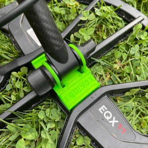 Puede incluir: Un detector de metales negro con un componente de plástico verde que dice "EQUINOX". El detector está sobre un lecho de hierba verde. El número de modelo "EQX 71" es visible en el detector.