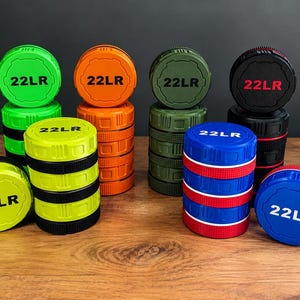 May include: A collection of cylindrical 22LR ammunition storage containers in various colors including neon green, orange, olive green, black, yellow, and blue. Each container features a textured grip and the text 22LR printed on the top lid.
