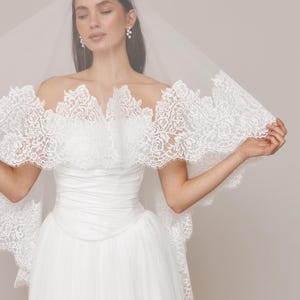 Op de afbeelding: Een witte trouwjurk met een tule rok en een strapless lijfje. De jurk heeft een delicate kanten rand rond de halslijn en mouwen. Een doorschijnende sluier bedekt het hoofd en de schouders.