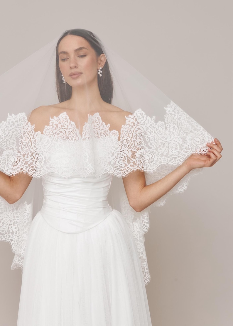 Op de afbeelding: Een witte trouwjurk met een tule rok en een strapless lijfje. De jurk heeft een delicate kanten rand rond de halslijn en mouwen. Een doorschijnende sluier bedekt het hoofd en de schouders.
