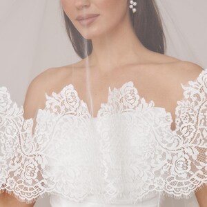 Op de afbeelding: Een bruid in een witte trouwjurk met een gedetailleerde kanten overlay. De jurk heeft een strapless ontwerp met bloemen kanten details. Een doorschijnende sluier is over de schouders gedrapeerd. Parel oorbellen maken de look af.