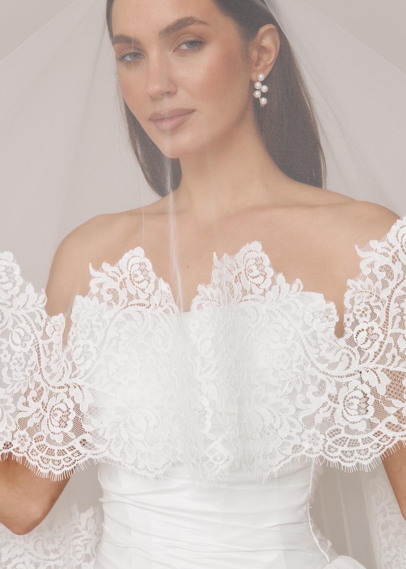 Op de afbeelding: Een bruid in een witte trouwjurk met een gedetailleerde kanten overlay. De jurk heeft een strapless ontwerp met bloemen kanten details. Een doorschijnende sluier is over de schouders gedrapeerd. Parel oorbellen maken de look af.
