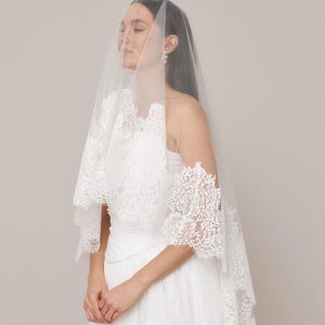Op de afbeelding: Een witte bruidssluier met ingewikkelde kanten details. De sluier is gedrapeerd over een witte jurk, wat een zachte, romantische uitstraling cre&euml;ert. De kanten rand voegt een delicate touch toe aan het bruidsensemble. De bruid draagt parel oorbellen.