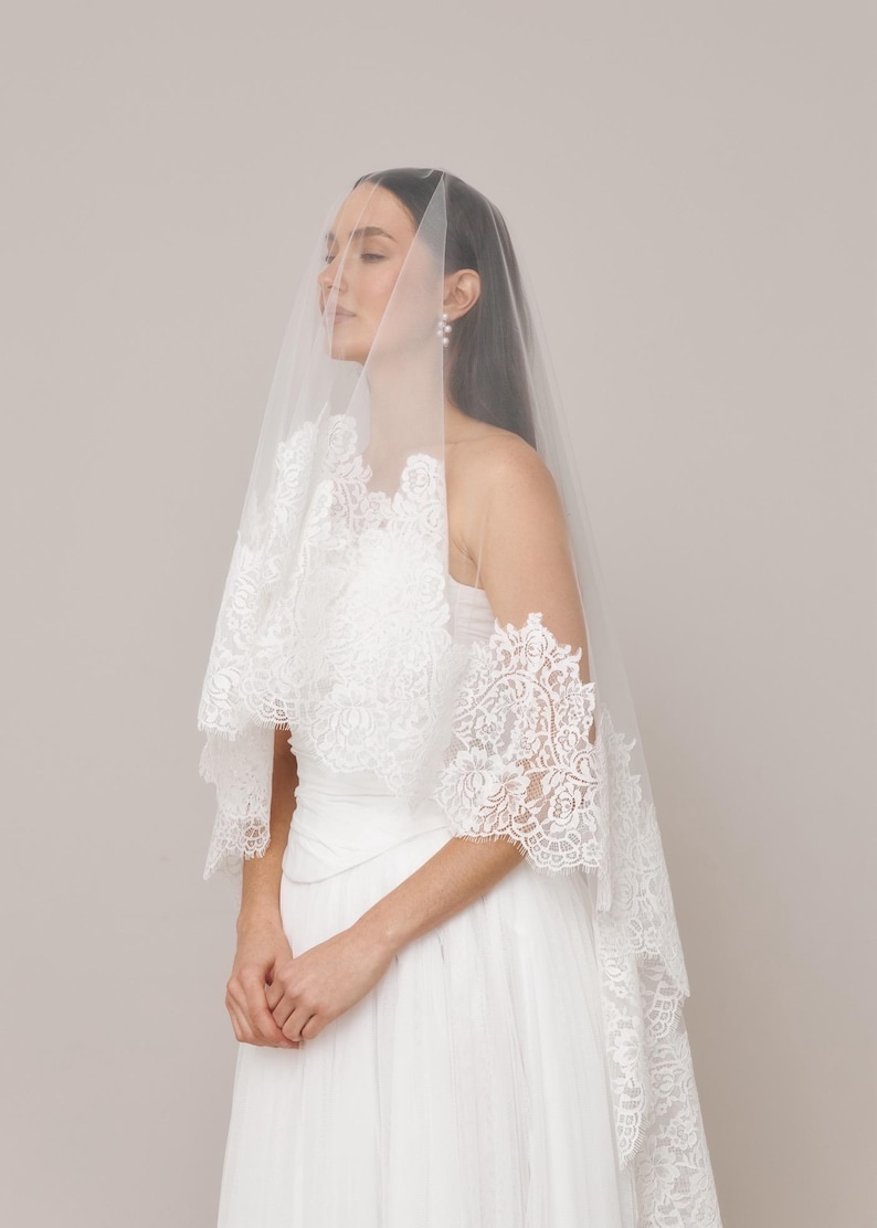 Op de afbeelding: Een witte bruidssluier met ingewikkelde kanten details. De sluier is gedrapeerd over een witte jurk, wat een zachte, romantische uitstraling cre&euml;ert. De kanten rand voegt een delicate touch toe aan het bruidsensemble. De bruid draagt parel oorbellen.