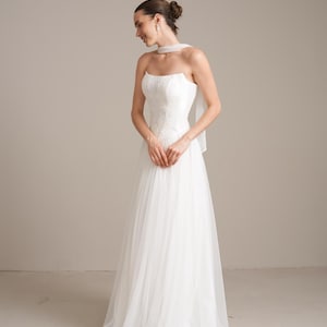May include: A floor-length white strapless gown featuring a fitted bodice with delicate lace detailing and a flowing tulle skirt. The dress includes a sheer fabric neck accessory that wraps around the neck and drapes down the back.