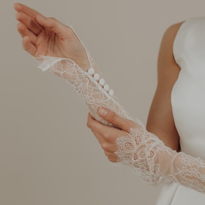 May include: White lace bridal gloves with button details. The gloves feature intricate floral lace patterns and are designed with a fitted silhouette. The buttons are small and round, adding a delicate touch to the design. The gloves are a wedding accessory.