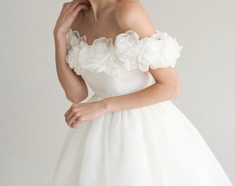 Op de afbeelding: Een witte off-the-shoulder jurk met bloemendetails. De jurk heeft een getailleerd lijfje en een uitlopende rok. De bloemversieringen zijn geconcentreerd rond de halslijn, wat een romantische en elegante uitstraling creëert. De jurk is geschikt voor speciale gelegenheden.