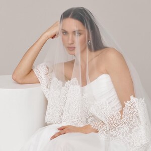 Op de afbeelding: Een bruid draagt een witte trouwjurk en sluier. De strapless jurk heeft een tule rok. De doorschijnende sluier bedekt het gezicht. De jurk heeft gedetailleerd kant op de mouwen en het lijfje, wat een elegante en romantische uitstraling cre&euml;ert.