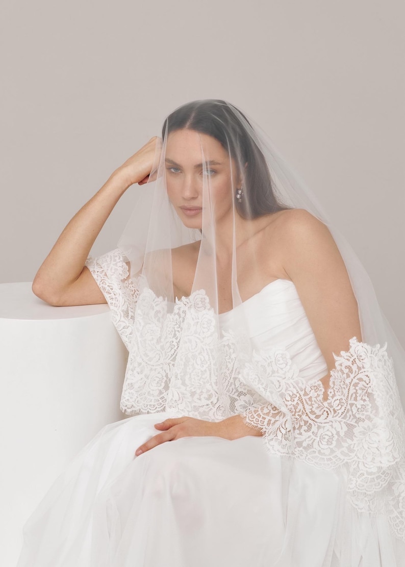Op de afbeelding: Een bruid draagt een witte trouwjurk en sluier. De strapless jurk heeft een tule rok. De doorschijnende sluier bedekt het gezicht. De jurk heeft gedetailleerd kant op de mouwen en het lijfje, wat een elegante en romantische uitstraling cre&euml;ert.