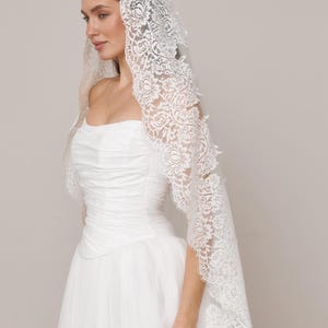 Op de afbeelding: Een witte bruidssluier met ingewikkelde bloemenkanten details. De sluier is gedrapeerd over een witte strapless trouwjurk. De kanten rand valt over de schouders en cre&euml;ert een elegante en romantische uitstraling.