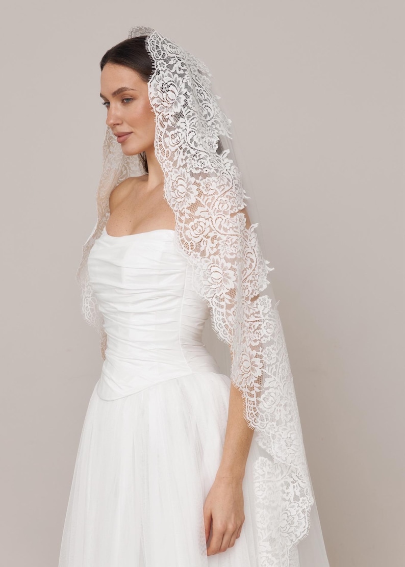 Op de afbeelding: Een witte bruidssluier met ingewikkelde bloemenkanten details. De sluier is gedrapeerd over een witte strapless trouwjurk. De kanten rand valt over de schouders en cre&euml;ert een elegante en romantische uitstraling.