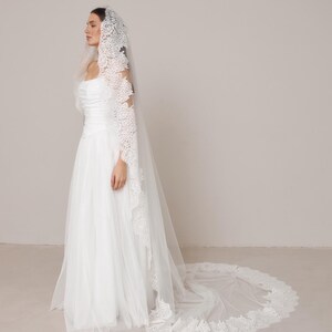 Op de afbeelding: Een witte trouwjurk met een lange, vloeiende tule rok en een bijpassende sluier. De sluier heeft ingewikkelde kanten details langs de randen en mouwen. De jurk heeft een getailleerd lijfje en een eenvoudig, elegant ontwerp.