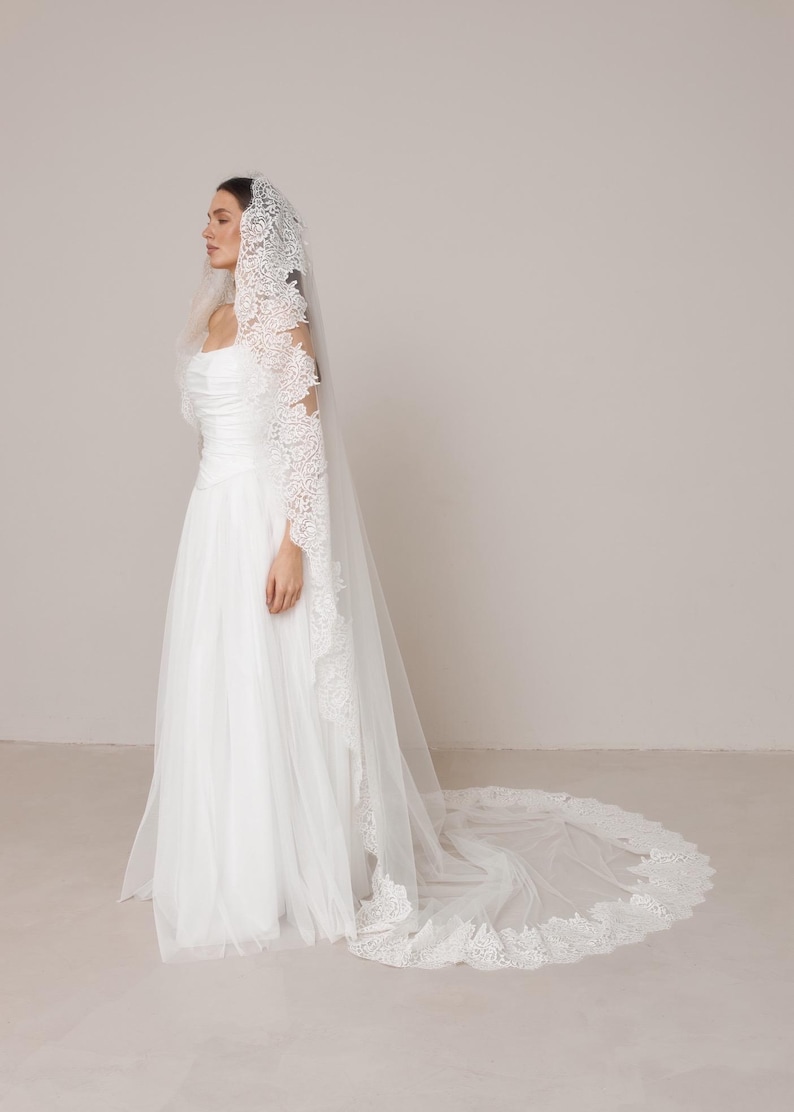 Op de afbeelding: Een witte trouwjurk met een lange, vloeiende tule rok en een bijpassende sluier. De sluier heeft ingewikkelde kanten details langs de randen en mouwen. De jurk heeft een getailleerd lijfje en een eenvoudig, elegant ontwerp.