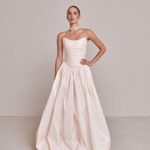 May include: A floor-length, strapless gown in a pale blush pink hue. The bodice features horizontal ruching and a structured, fitted shape, while the skirt is voluminous with deep pleats, creating a full, gathered silhouette that reaches the floor.