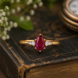 May include: A gold ring with a central, pear-cut, deep red gemstone, and smaller, round, clear stones. The ring is set on a vintage, leather-bound book, with a blurred background of foliage and a pocket watch.