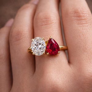 May include: A gold ring featuring two stones set side-by-side: a clear, oval-cut gemstone and a vibrant, pear-shaped red ruby. The stones are held in place by gold prongs on a polished gold band, creating a distinct two-stone design on the finger.