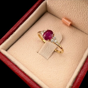 May include: A gold ring with a large oval ruby gemstone and small accent diamonds. The ring is displayed in a red jewelry box with a beige interior. The ruby is a deep red color, and the diamonds sparkle.