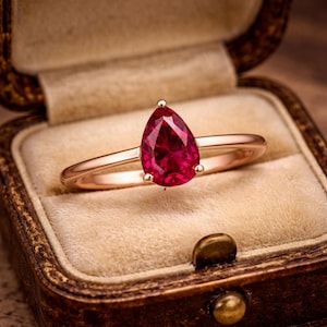 Pode incluir: Um anel com uma pedra preciosa vermelho-profundo em forma de pera, montada numa banda de ouro rosa polido. O anel apresenta uma pedra facetada central segura por garras, exibindo um tom carmesim vibrante e um acabamento metálico liso.