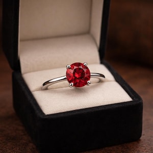 Può includere: Un anello d'argento con una grande gemma rossa rotonda, presentato in un portagioie di velluto nero con interno color crema. La fascia dell'anello è sottile e lucida.