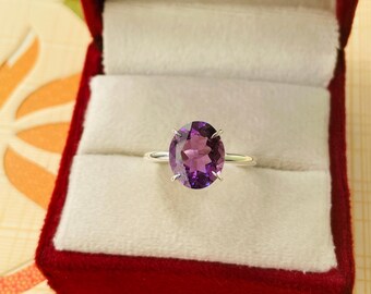 May include: A silver ring with a large, oval-cut amethyst gemstone. The ring is set in a classic four-prong setting and is displayed in a red velvet ring box. The amethyst has a deep purple hue.