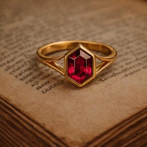 May include: A gold ring featuring a large, faceted, red gemstone in a hexagonal setting. The ring's band has a split design. The ring is set on a vintage book page.