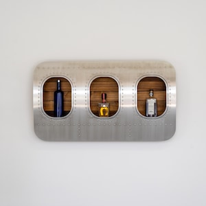 May include: A wall-mounted liquor shelf with three oval openings. The shelf is made of brushed metal with rivets. Each opening has a wooden backing and holds a different bottle of liquor. The bottles are blue, amber, and clear.