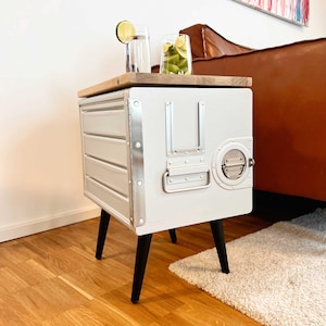 Può includere: Un tavolino da salotto realizzato con un carrello di servizio aereo in metallo grigio chiaro riutilizzato e un piano in legno naturale. L'unità presenta un corpo rettangolare con nervature orizzontali, una maniglia in metallo argentato e quattro gambe nere.