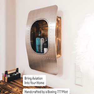 May include: A wall-mounted cabinet made from a piece of aircraft, with a wooden interior and a metal exterior. The cabinet has an oval opening and holds two bottles. The text "Bring Aviation Into Your Home" and "Handcrafted by a Boeing 777 Pilot" are visible.