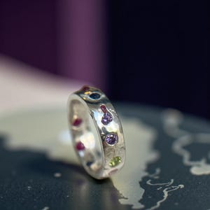 Puede incluir: Un anillo de plata con una superficie irregular, adornado con piedras preciosas de varios colores. El anillo presenta una mezcla de piedras moradas, rojas, verdes y azules, creando un diseño único. Es una joya.