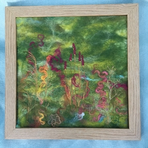 May include: A framed needle felted artwork depicting a green landscape with colorful flowers and foliage. The artwork is framed in a light wood frame.