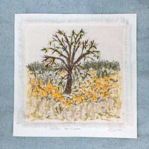 Könnte beinhalten: Eine gestickte Kunstarbeit, die einen gelben blühenden Ginsterbusch mit einem kleinen Baum in der Mitte zeigt. Das Kunstwerk ist mit einem weißen Rahmen versehen und trägt den Text "gorse in bloom" und "Alecia" unterhalb des Bildes.
