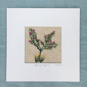 Könnte beinhalten: Eine gerahmte Stickerei eines rosa Magnolienbaums mit gelben Blüten an der Basis. Die Stickerei ist auf einem beigefarbenen Stoff und trägt den Titel "Spring Magnolia" mit dem Namen des Künstlers "Aleason" darunter.