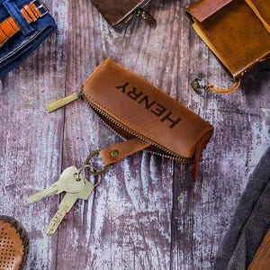 Puede incluir: Una vista aérea de accesorios para hombres, que incluyen un llavero de cuero marrón con "HENRY" impreso, un reloj con una correa de cuero naranja, una cartera de cuero marrón, un diario y zapatos de cuero marrón. Los artículos están dispuestos sobre una superficie de madera.