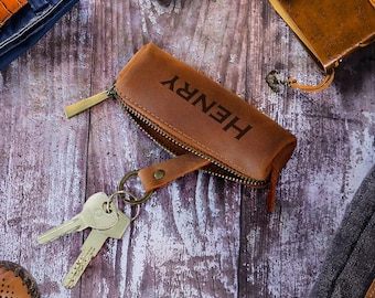 May include: An overhead shot of men's accessories, including a brown leather key case with "HENRY" printed on it, a watch with an orange leather band, a brown leather wallet, a journal, and a pair of brown leather shoes. The items are arranged on a wooden surface.