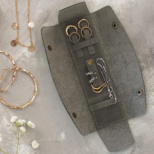 Può includere: Un assortimento di gioielli dorati, tra cui orecchini a cerchio, orecchini a lobo, collane e anelli, è esposto su una superficie grigia. Un astuccio portagioie in pelle grigia è aperto, mostrando anelli e orecchini aggiuntivi. Fiori bianchi aggiungono un tocco delicato.