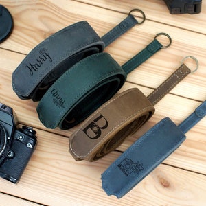 May include: Three personalized leather camera straps in green, brown, and gray with the names "Harry", "Anna", and "B" engraved on them. A fourth strap is blue with a camera icon and the word "Melissa" engraved on it.