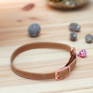 May include: A brown leather cat collar with a rose gold buckle and a pink bell.