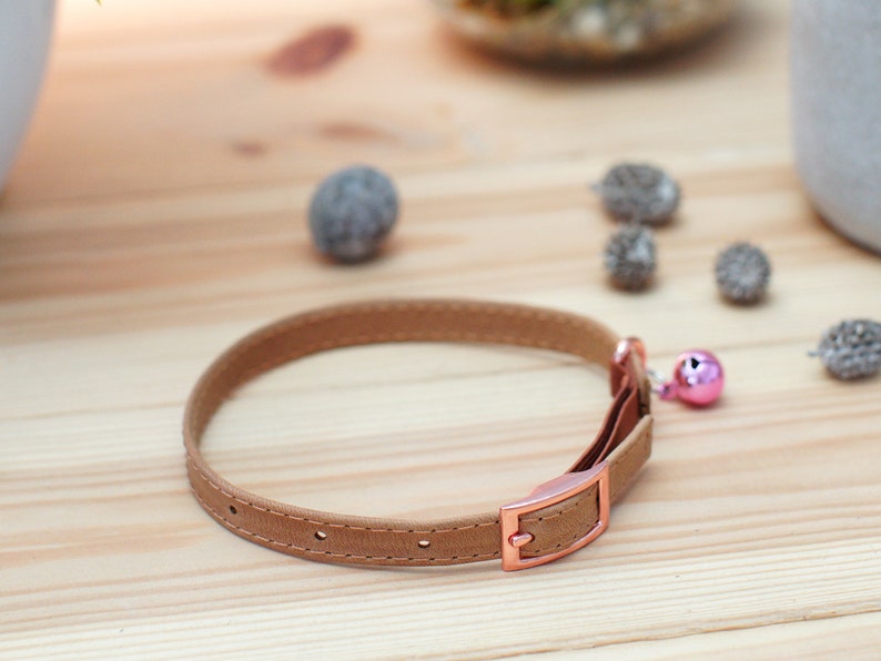 May include: A brown leather cat collar with a rose gold buckle and a pink bell.