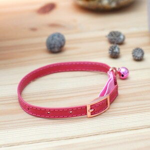 May include: A pink leather cat collar with a pink ribbon and a gold buckle. The collar has a small pink bell attached.