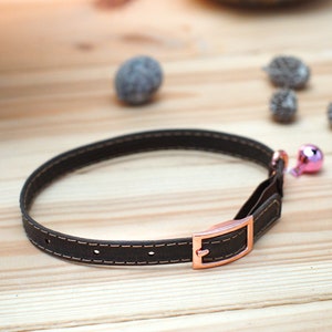 May include: A brown leather cat collar with a rose gold buckle and a pink bell.