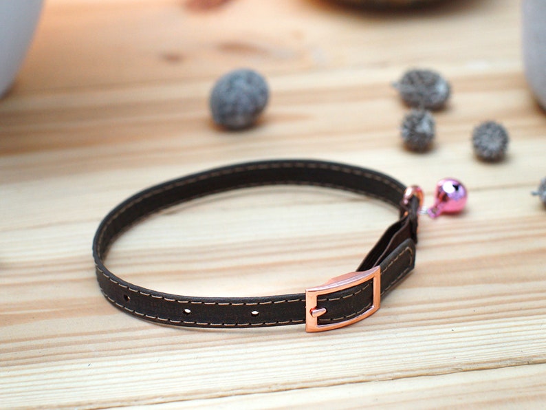 May include: A brown leather cat collar with a rose gold buckle and a pink bell.